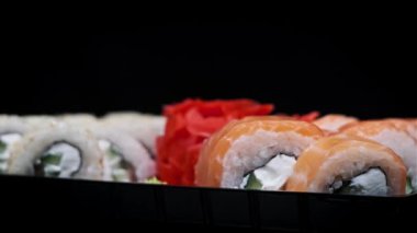 Suşi plastik bir kutuda yuvarlanır ve siyah arka planda yakın plan çekilir. Hazır suşi kutusu ve farklı lezzetli Japon dürümleri bir teslimat konteynerinde. Yemek teslimatı, paket yemek.