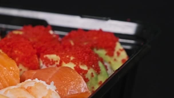 プラスチック製の箱に入った寿司は 黒い背景のクローズアップを回転させる デリバリーコンテナに異なる美味しい日本ロールのセット寿司ボックス 食糧配達 テイクアウト食品のコンセプト — ストック動画