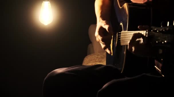 暖かい光で暗い部屋でアコースティックギターを演奏する若者 ミュージシャンは 単一の光源によって照らされ 劇的な影を投げかけます 薄暗い光でギタリスト 歌を演奏することを学ぶ ライフスタイル — ストック動画