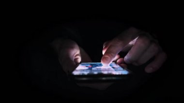 Karanlıkta akıllı telefon kullanarak ekranın mavi ışığını parlaklaştıran ellerin yakın çekimi. Adam sosyal medya haberlerini karanlık bir odada geniş açılı bir akıllı telefondan aktarıyor.
