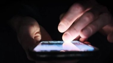 Erkek el, sosyal medya haberlerini karanlık bir odada, geniş açılı, yakın plan bir telefondan aktarıyor. Tamamen karanlıkta akıllı bir telefon kullanan kişi, parlayan ekrana ve parmaklarına odaklanmış..
