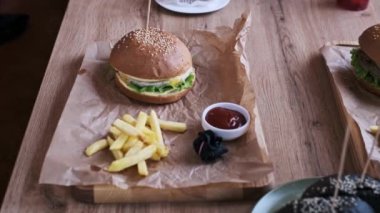 İki burger, biri klasik, biri siyah çörekli, bir kafe masasında patates kızartması ve ketçapın yanında sunuluyor. Sahne, lezzetli ve sosyal bir yemek deneyimini yakalar..