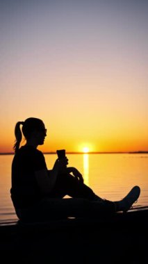 Batan güneşin sıcacık parıltısıyla yıkanan genç bir kadın, sakin gölün kenarında bir fincan kahvenin tadını çıkarırken teselli ve huzur buluyor. Gökyüzünün büyüleyici renkleri resim gibi bir zemin oluşturur..