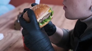 Siyah eldiven giyen bir kadın sulu bir burgerden büyük bir ısırık alır. Yakın çekim, fast food ile ilişkili tüketim ve hoşgörü eylemini vurguluyor ve sağlık ve gıda üzerinde bir yansımaya yol açıyor.