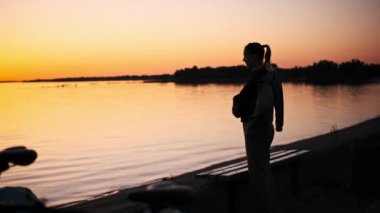 Bir kadın gün batımında gölün kenarında duruyor. Rengarenk gökyüzüne yaslanmış. Rahatlatıcı bir aradan sonra ceketini giyiyor..