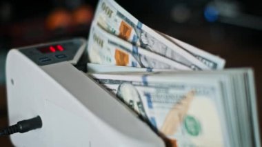 Bir para sayma makinesi bir yığın Amerikan doları banknotunu hızlı bir şekilde işliyor ve toplamı verimli bir şekilde hesaplıyor. Hışırtı banknotlarının gıcır gıcır sesi zenginlik ve finansal refahı çağrıştırıyor..