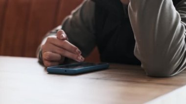 Bir kadın akıllı telefonunu parmağıyla yavaşça kaydırarak kullanır. Mavi telefon bir kafede sıcak tonlu ahşap bir masanın üzerinde duruyor. 50 fps 'de pürüzsüz ve detaylı bir video oluşturuyor..