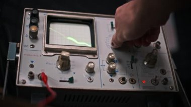 Bir el, gösterilen yeşil dalga şeklini değiştirerek eski bir osiloskopun düğmelerini manipüle ediyor. Bu görüntü analog sinyal analizindeki hassasiyeti ve kontrolü yakalar..