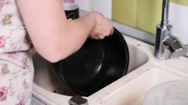 Bir kadın ağır yanmış bir tencereyi mutfak lavabosunda duruluyor ve temizliğe hazırlıyor. Esrar önemli miktarda kullanım ve yanmış gıda kalıntısı gösteriyor..