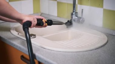 Bir kadın derin bir mutfak lavabosunu temizler. Elle tutulan bir buhar temizleyicisi ve fırça ile. Yüzeydeki kiri ve bakterileri temizlemeye odaklanır..
