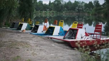 Sakin bir gölün kıyısında renkli pedallı gemiler yolcuları bekliyor. Neşeli renkleri ve yemyeşil ağaçları mükemmel bir yaz günü için sakin bir zemin oluşturur..