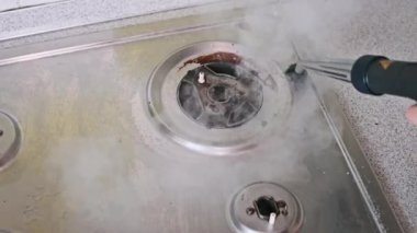 Yakın çekim, yağlı bir gaz ocağını titizlikle temizlemek için kullanılan yuvarlak fırçalı bir buhar temizleyiciyi gösteriyor. Buhar kiri ve kiri etkili bir şekilde eritiyor ve lekesiz bir son ortaya çıkıyor.