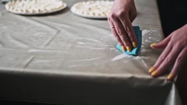 Sarı tırnaklı kadın, yemek hazırladıktan sonra gri masa örtüsünden un temizlemek için mavi bez kullanıyor. Mutfak hijyeninin önemini vurguluyor..