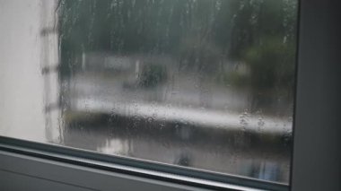 Yağmurlu bir günde bu atmosferik 4K görüntüleriyle şehri inceleyin. Pencereden süzülen yağmur damlaları, binaların ve ağaçların görüntüsünü bulanıklaştırıyor, huzur ve yalnızlık hissi yaratıyor..