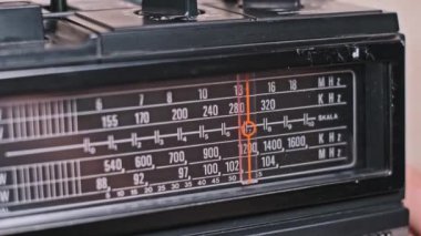 Klasik radyo kadranını ayarlayan bir elin yakın çekimi, frekans bandında hareket eden bir iğne. Görüntü, analog radyo günlerinde bir istasyon bulmanın kesin eylemini yakalar..