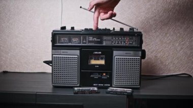Bir parmak çalma tuşuna basar, sonra klasik bir teybin durdurma tuşuna basar, analog sesi kontrol etmenin tatmin edici dokunsal deneyimini vurgular. Müzik tutkunları için nostaljik bir imaj.