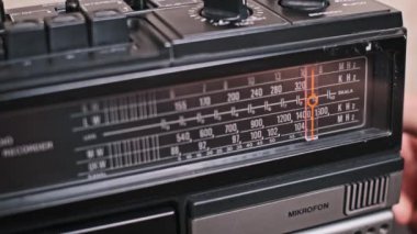 Radyoda ayarlanan klasik bir teybin yakın plan görüntüsü. Bir el düğmeyi ayarlar, FM bandında bir istasyon arar, beklenti ve analog teknolojinin cazibesini uyandırır..