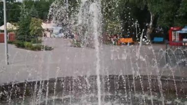 Canlı şehir meydanı manzarası serinletici bir çeşme ile merkez olarak, hareketli etkinlik ve yaz titreşimleriyle çevrili.