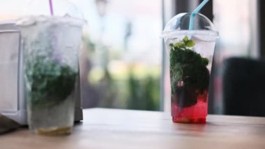 Kafedeki bir masada plastik bardakta iki ferahlatıcı mojito kokteyli. Naneli, limonlu ve buzlu yaz içkileri, arka planda bulanık sokak manzarası.