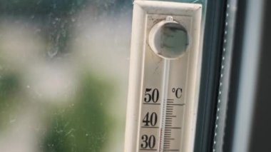 Pencereye bağlı bir termometrenin yakın çekimi, yüksek sıcaklığın 40 dereceyi aştığını gösteriyor. Arka plandaki bulanık yeşil yapraklar bunaltıcı bir yaz günü olduğunu gösteriyor..