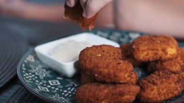 Bir tabak altın-kahverengi tavuk nugget 'ına yakın plan, yanında da küçük bir kase kremalı beyaz sos. Bir el nuggetı daldırmak için uzanır, çıtır kıtır dokular ve lezzetin mükemmel bir kombinasyonu.