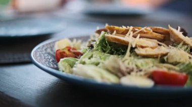 Sezar salatasını yakın plan bir tabakta, bir restorandaki ahşap bir masanın üzerine koyun. Dilimlenmiş tavuk göğsü, kiraz domatesleri, marul yaprakları, parmesan peyniri, sos lezzetli bir yemek deneyimi yaratır.