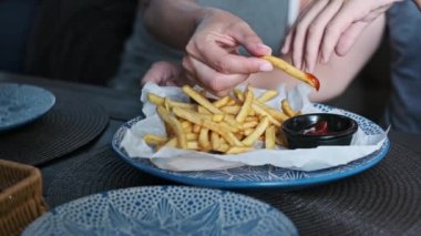 Yakın çekim, bir elin çıtır patatesleri kırmızı sosa batırdığı anı yakalar. Gıdanın dokularına ve renklerine odaklanmak hoşgörü ve tatmin duygusunu çağrıştırıyor..