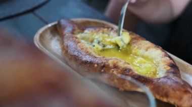 Erimiş peynirin içine yumurta sarısı karıştırmak için çatal kullanan bir elin yakın plan fotoğrafı. Yeni pişmiş Adjarian Khachapuri 'de görsel olarak çekici ve iştah açıcı bir sahne yaratıyor..