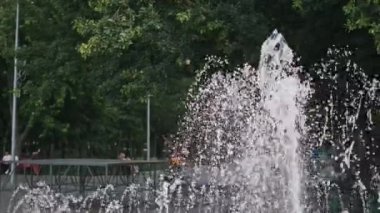 Şehir parkı çeşmesinde su jetleri dans ediyor ve ışıldıyor. Sıcak bir yaz gününde ferahlatıcı bir kaçış sunuyor. Arka plandaki manzaranın keyfini çıkarıyorlar..