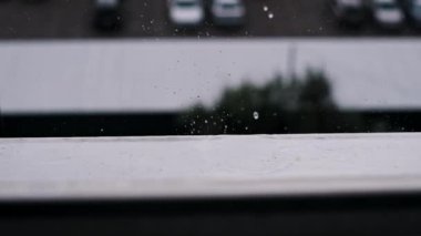 Pencerenin pervazına düşen yağmur damlalarının yakın çekim görüntüleri. Otopark ve binaların bulanık bir görüntüsü var. Yalnızlık ve tefekkür hissi veriyor..
