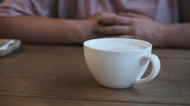 Bir bardak kapuçino ve latte sanatı sizi bir yudum içmeye ve anın tadını çıkarmaya davet eden ahşap bir masada otururlar..