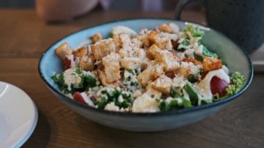 Taze ve lezzetli bir Sezar salatası, kıtır kıtır ekmek, parmesan peyniri ve sulu domatesli, kırsal ahşap bir masada mavi bir kasede servis edilir, iştah açıcı bir sahne yaratır..
