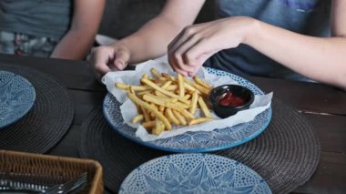 Bir çocuğun eli çıtır çıtır bir patates kızartmasına uzanır ve onu nazikçe küçük bir kase ketçapa batırır. Harekete odaklanma, klasik bir fast food yemeğinin tadını çıkarmanın basit zevkini vurgular..