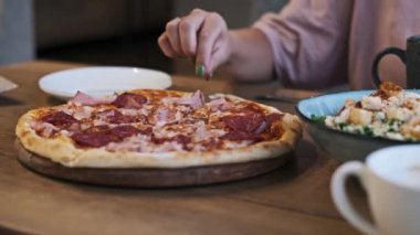 Yeşil ojeli kadın eli, salam ve pastırma ile lezzetli bir pizzayı kesiyor. Arka plandaki Sezar salatası ve kahve kafe yemek sahnesini tamamlıyor..