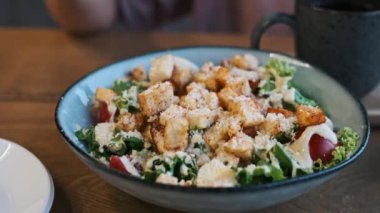 Tavuklu Sezar salatası, marul, kiraz domatesleri, kızarmış ekmek ve kafedeki ahşap bir masada parmesan peyniri. Arka planda, telefonlu bulanık bir kadın eli silueti var..