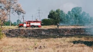 İtfaiyeciler, arka planda bir itfaiye aracıyla bir yangını söndürmek için çalışıyor. Yangın tehlikelerini ve yangın güvenliğinin önemini vurguluyorlar..