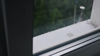 Pencerenin pervazına yakın çekim, yağmur damlaları dışarı düşüyor, yemyeşil manzarayı bulanıklaştırıyor. Sağanak yağış sırasında doğanın güzelliğini ve huzurunu çağrıştırıyor..