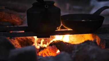 Bir kadın eli, arka planda kaynayan bir çaydanlıkla, canlı bir kamp ateşinin üzerinde tavuğun parçalarını ustalıkla karıştırır. Açık hava yemekleri ve maceranın kırsal bir sahnesi..