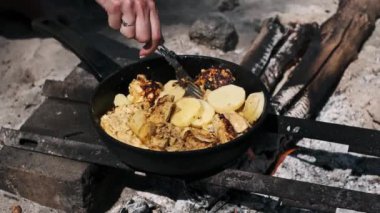 Bir kadın eli, tavuğu ve patatesleri karıştırmak için çatal kullanıyor. Kamp ateşinin üstünde demir tavada kızartıyorlar..