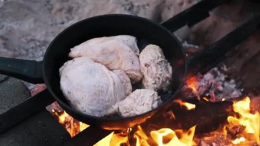 Kamp ateşinin tepsisinde çiğ tavuk parçaları. Alevler kenarları yalıyor. Sahne, doğal ortamda basit ve otantik yemekleri temsil ediyor..