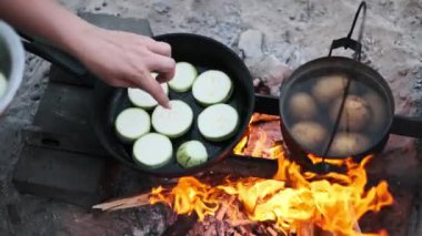 Bir el, kabağı bir kamp ateşinin üzerinde tavaya dilimliyor. Yanında bir tencere patates kaynıyor ve kırsal alanda pişirme sergiliyor..