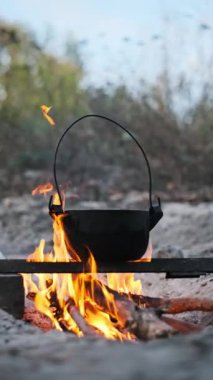 Siyah metal bir tencere alevli bir kamp ateşinin üzerinde asılı, vahşi doğada yemek pişiriyor. Alevler tencerenin dibini yalayarak sıcak ve davetkar bir atmosfer yaratıyor..