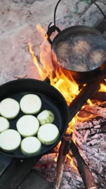 Kamp ateşinde tavada kızaran kabak dilimlerinin dikey videosu. Arka planda patates kaynıyor. Lezzetli ve sağlıklı bir yemek doğada hazırlanıyor..