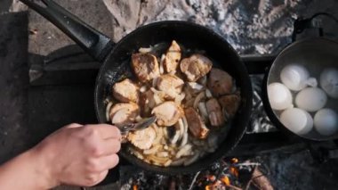 Bir el, kamp ateşinde tavada kızaran tavuk ve soğan parçalarını karıştırıyor. Yanında bir tencere haşlanmış yumurta var, yemeği tamamlamaya hazır..