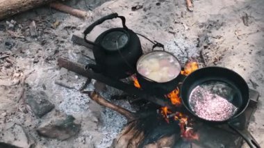 Bir çaydanlık, bir tencere güveç ve yağlı tavayla cızırdayan bir kamp ateşinin tepeden aşağı görüntüsü..