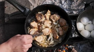 Bir insan, kamp ateşinde tavada kızartılmış soğan ve tavuk parçaları karıştırır. Yumurtalar yakındaki bir tencerede kaynamış, kırsal alanda pişirme sahnesi yaratıyor..
