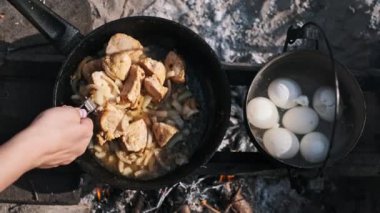 Bir el, kamp ateşinin üstünde tavukla soğan karıştırıyor. Arka planda bir tencere yumurta kaynıyor..