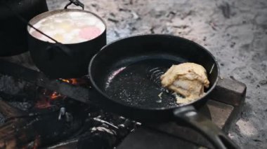 Kamp ateşinin üstünde et pişirmek, yakınlarda kaynayan bir çorba tenceresi, kırsal bir yemek pişirme deneyimini temsil ediyor..