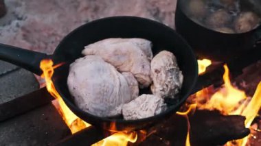 Kızartma tavası, pişmemiş tavuk ve bir tencere haşlanmış patatesle bir kamp ateşinin üzerinde oturuyor. Sahnede kırsal bir açık hava yemeği hazırlanıyor..