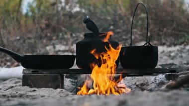 Kükreyen bir kamp ateşi, siyah bir çaydanlık, tencere ve kızartma tavası..